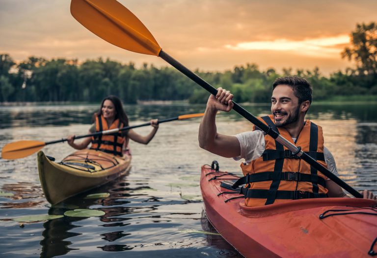 Ein lächelnder Mann und eine Frau paddeln in ihren Kajaks auf einem ruhigen See. Die warme Abendsonne taucht die Szene in ein stimmungsvolles Licht und unterstreicht das entspannte Naturerlebnis.